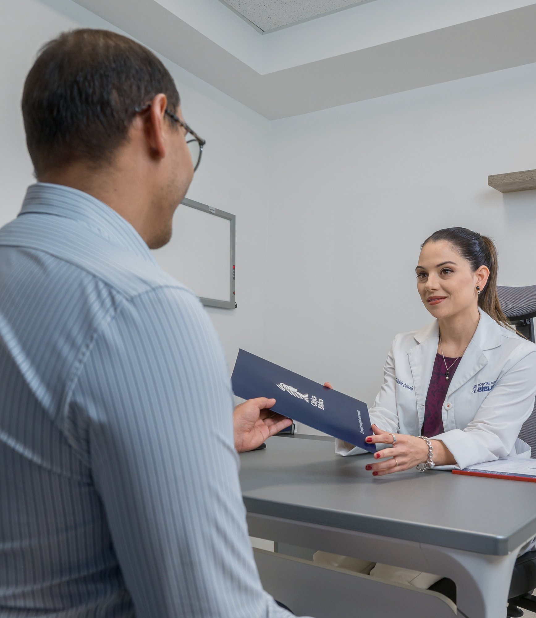 Doctor handing out a Clínica Bíblica information brochure to a patient during a medical consultation.
