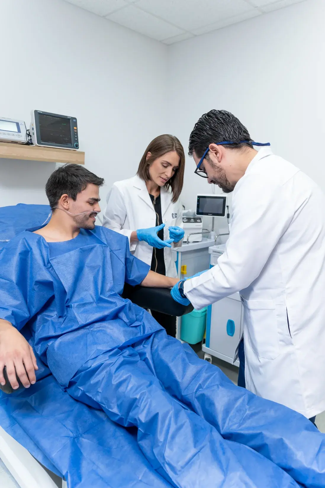 Paciente con bata azul recibe atención médica de dos profesionales de salud en una sala de procedimientos.