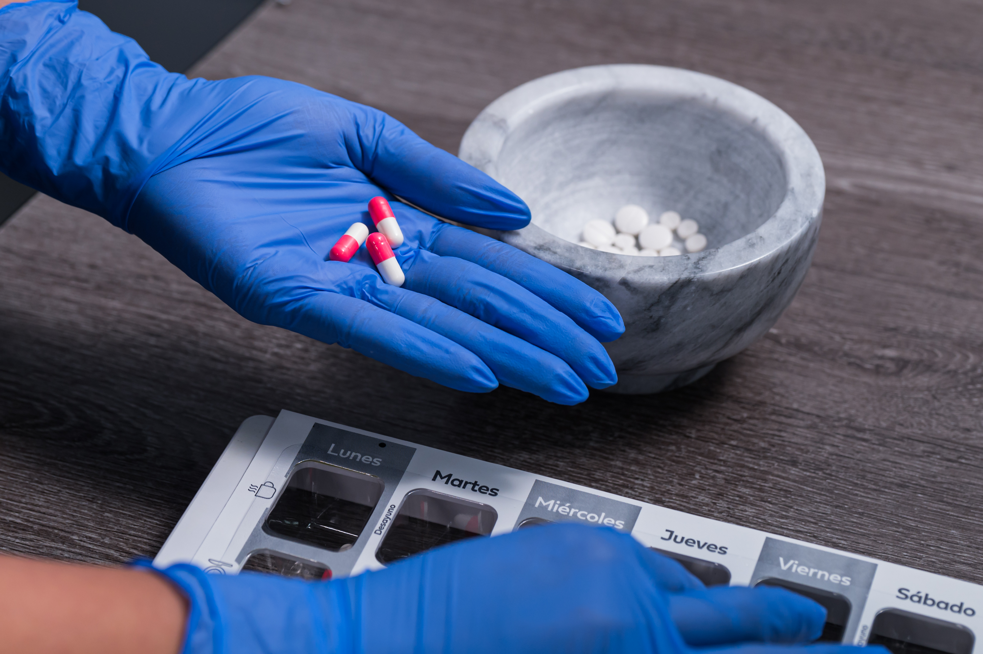 Hand with blue gloves holding red and white capsules next to a mortar with tablets and a weekly medication organizer.