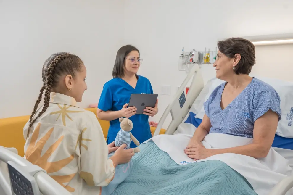 Enfermera hablando con paciente hospitalizada y una niña que sostiene un muñeco en una habitación médica luminosa.