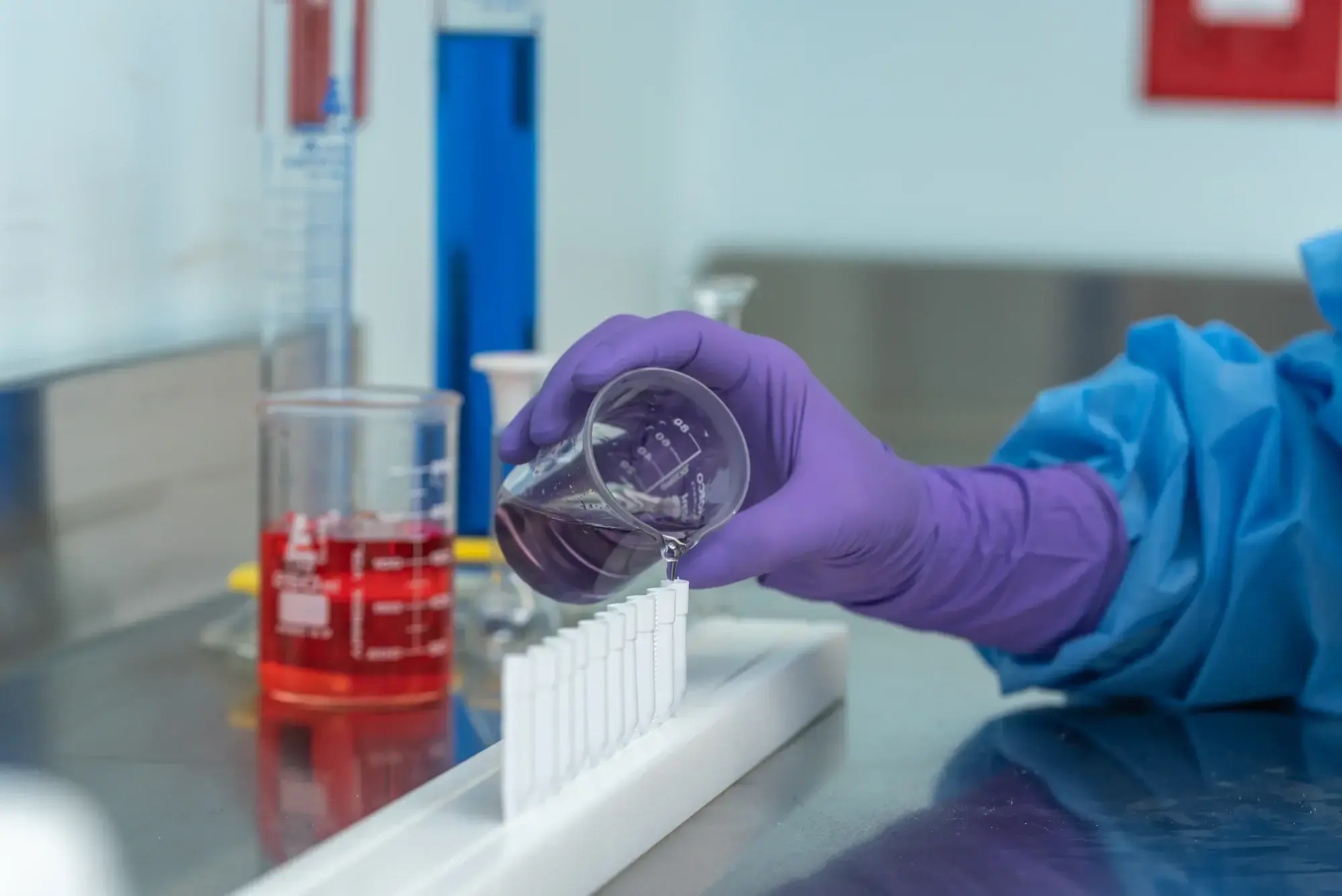 Mano con guante morado vertiendo solución púrpura en tubos de ensayo alineados en laboratorio.